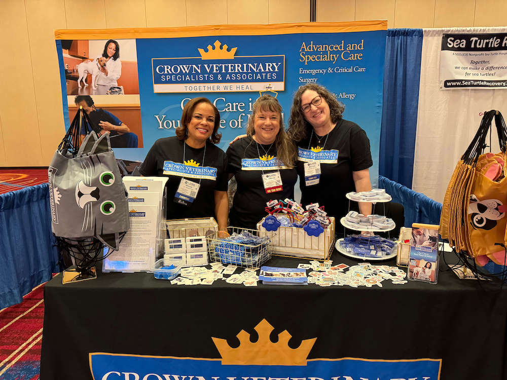 Three people smiling behind a veterinary booth with promotional items and signage for Crown Veterinary Specialists & Associates, at an indoor event space.